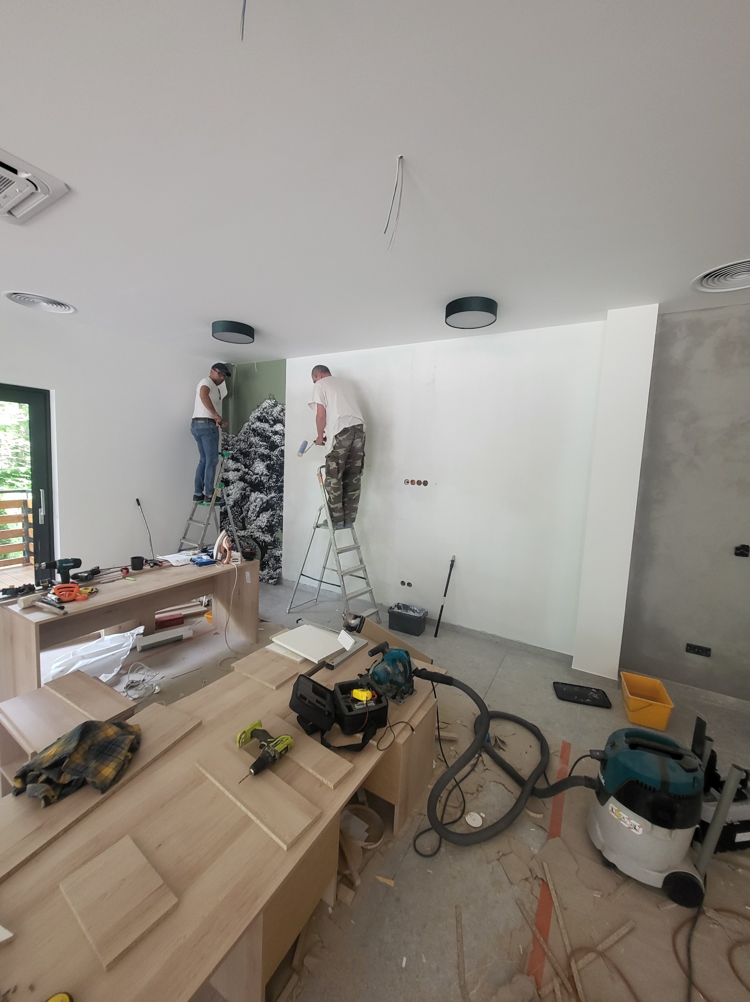 Dwóch mężczyzn na drabinach podczas remontu wnętrza, jeden maluje ścianę, drugi kładzie tapetę z motywem drzew, widoczne narzędzia i przygotowane meble.