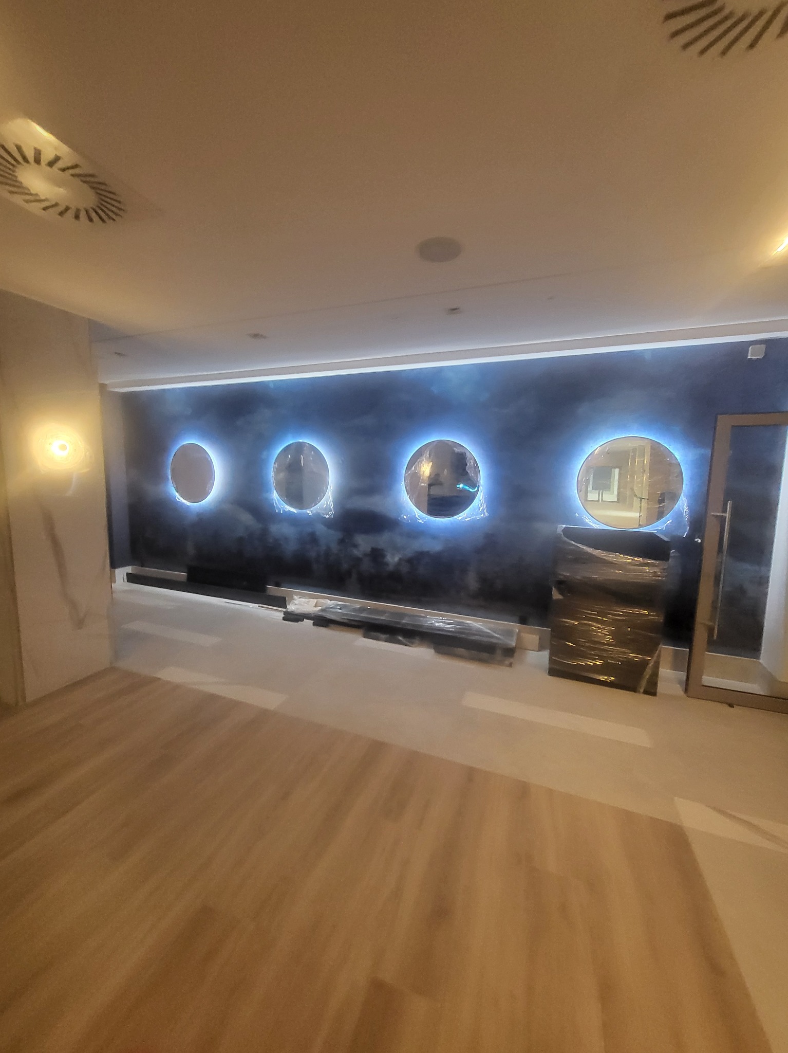 Ściana z efektem ombre w odcieniach granatu i szarości, ozdobiona czterema podświetlanymi okrągłymi dekoracjami, częściowo zabezpieczona folią ochronną, w trakcie prac wykończeniowych.