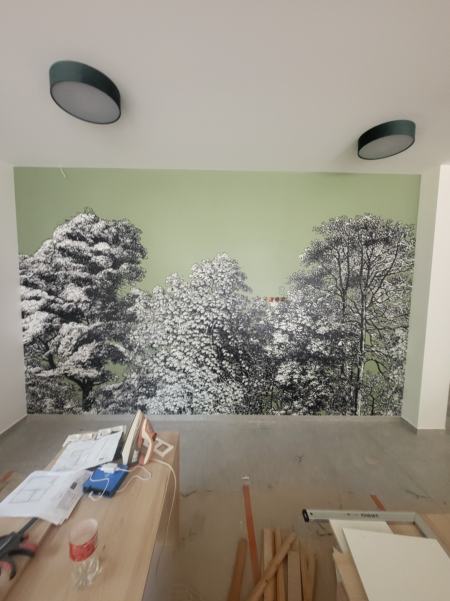 Ściana z zielonym tłem i czarno-białym motywem drzew, widoczne narzędzia i materiały na stole oraz podłodze podczas prac wykończeniowych.