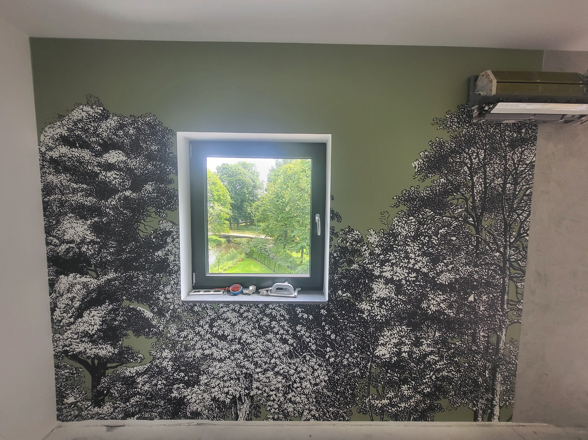 Pokój z zielonymi ścianami ozdobionymi tapetą z motywem drzew, widok z okna na park, narzędzia na parapecie, klimatyzator na górze ściany.