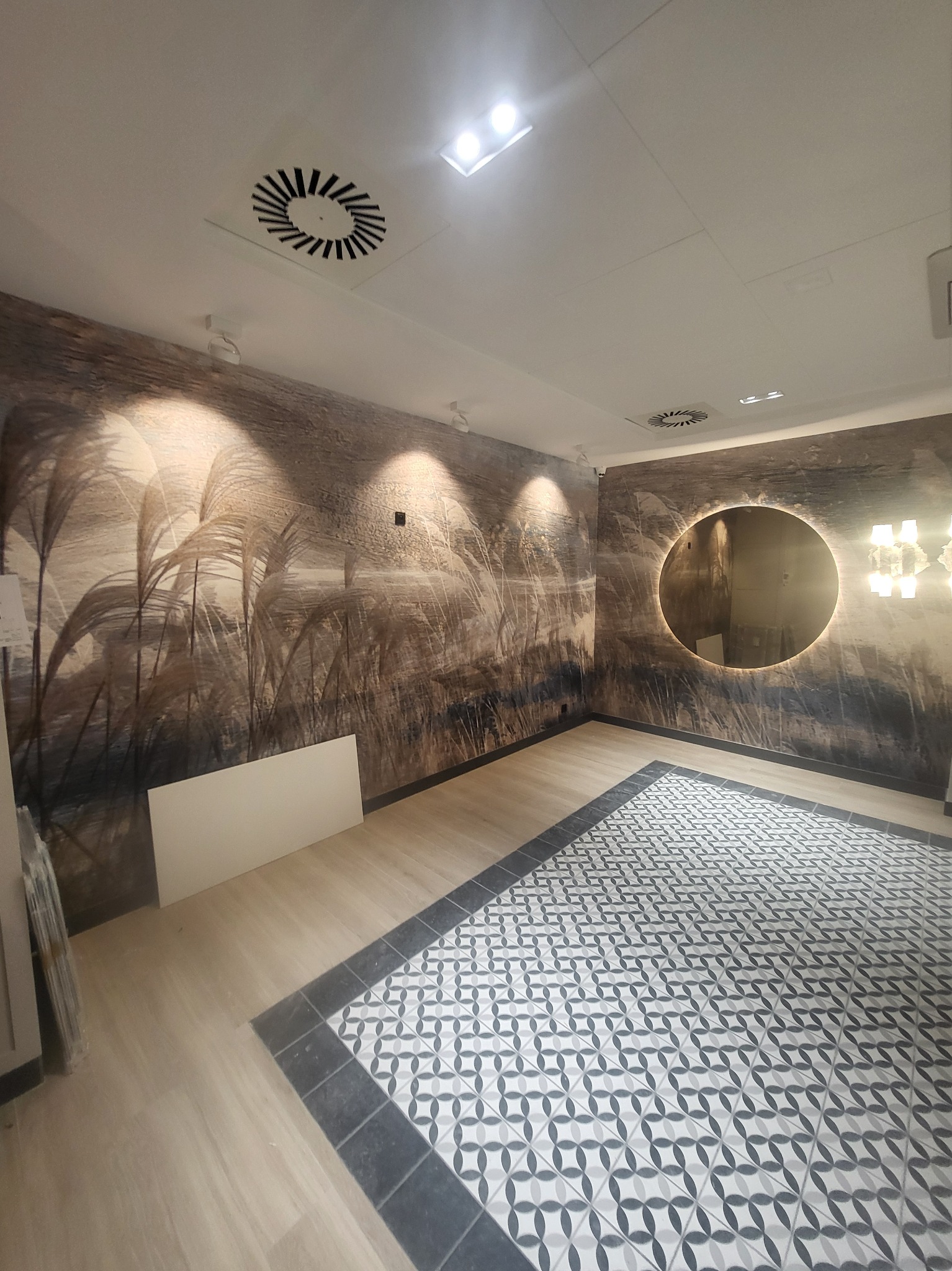 Wnętrze po remoncie z fototapetą z motywem traw, okrągłym lustrem z podświetleniem i wzorzystą podłogą, widoczne panele podłogowe i białe płyty na ścianie.