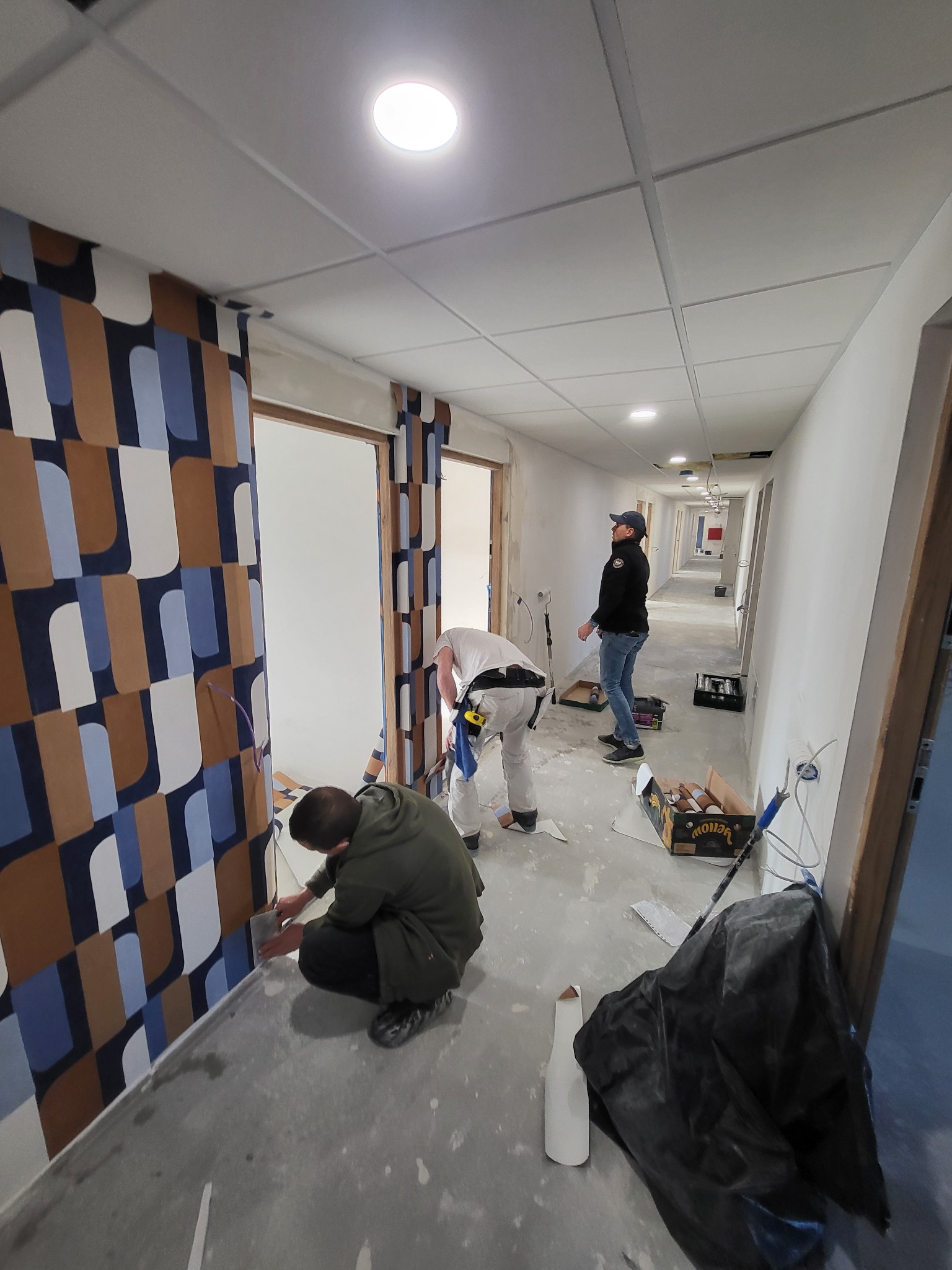 Montaż wzorzystej tapety w korytarzu biurowym; ekipa remontowa w trakcie pracy, widoczne narzędzia i niedokończone fragmenty ściany.