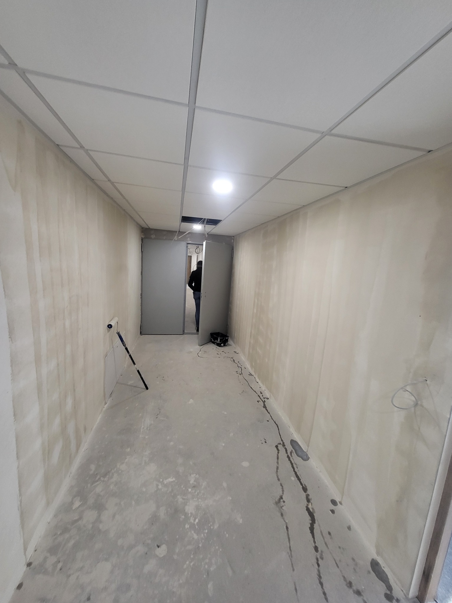 Korytarz w trakcie prac wykończeniowych: ściany pokryte wstępną warstwą, widoczne narzędzia malarskie, osoba w tle przechodząca przez drzwi.