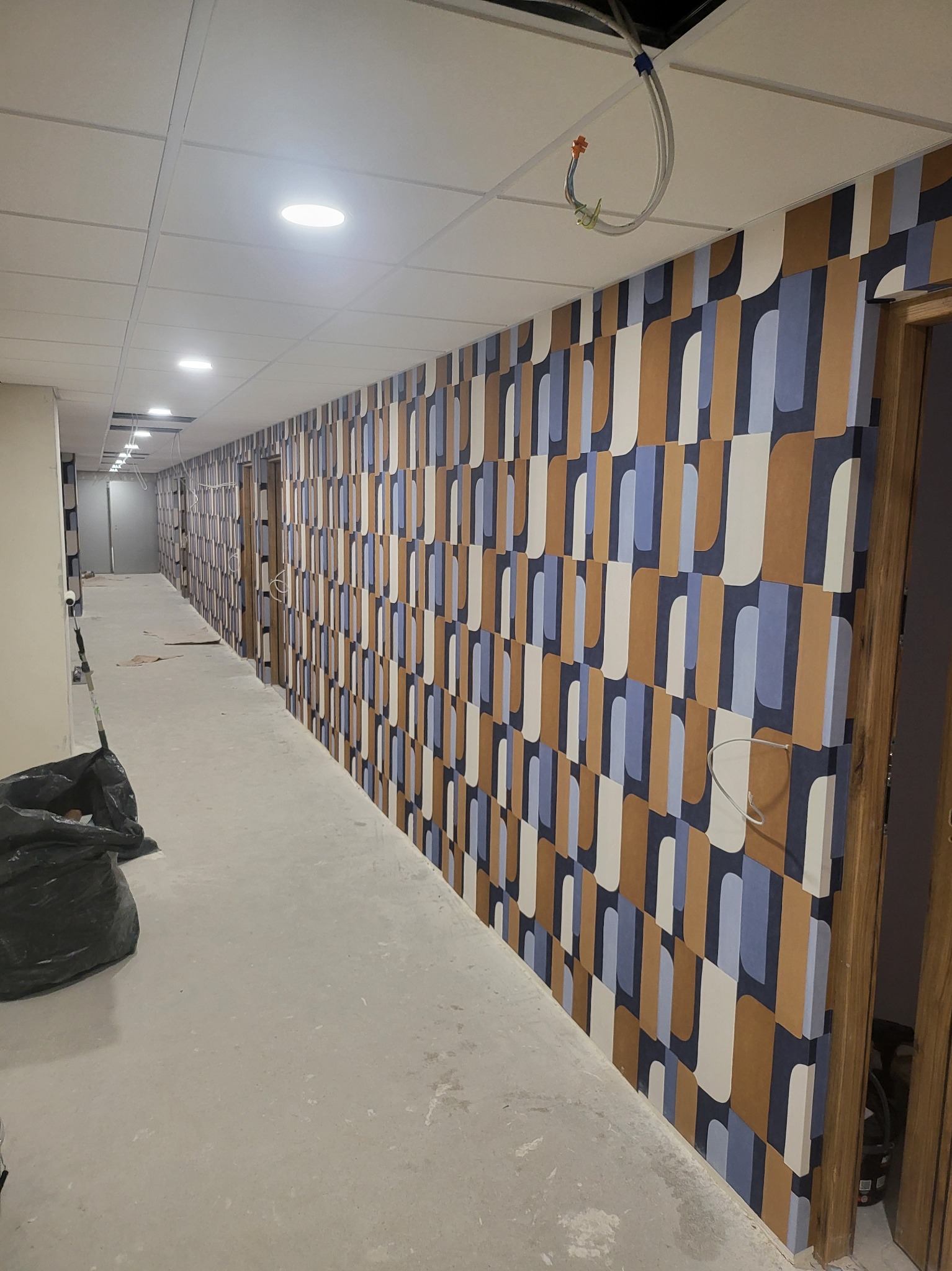 Długi korytarz w trakcie remontu, ściany pokryte tapetą w geometryczne wzory w odcieniach brązu, błękitu i bieli, częściowo zakryte kable wystające z sufitu, podłoga zabezpieczona folią malarską...