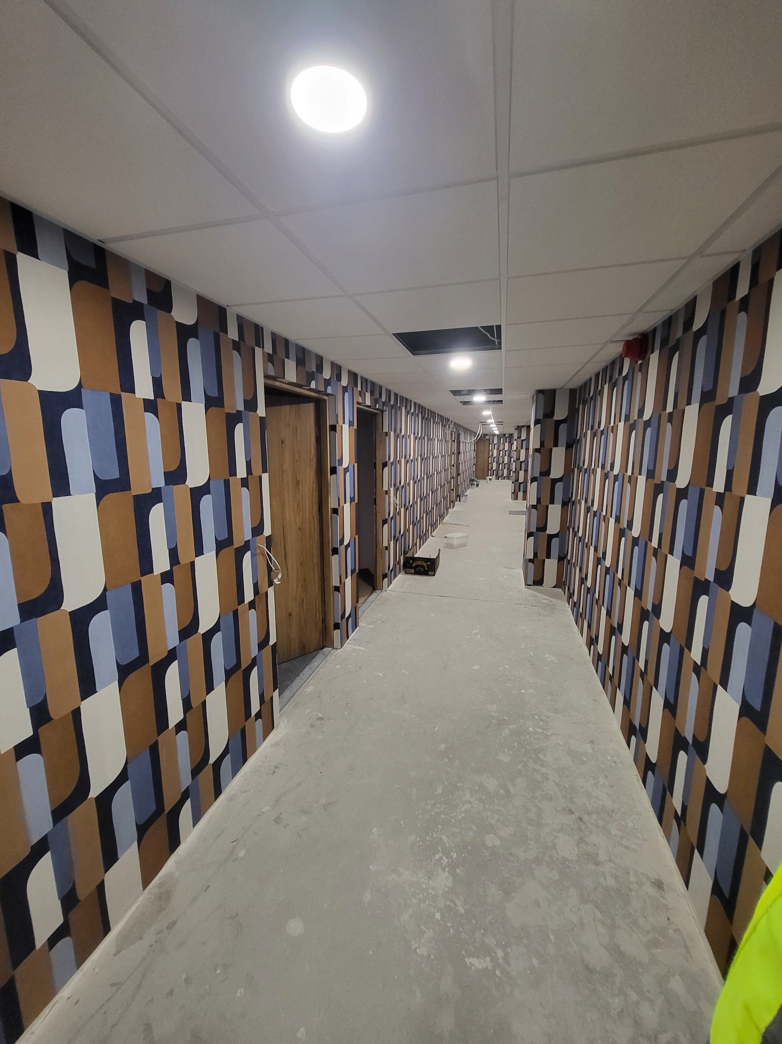 Długi korytarz biurowy po tapetowaniu ścian w geometryczny wzór, podłoga w trakcie prac wykończeniowych.