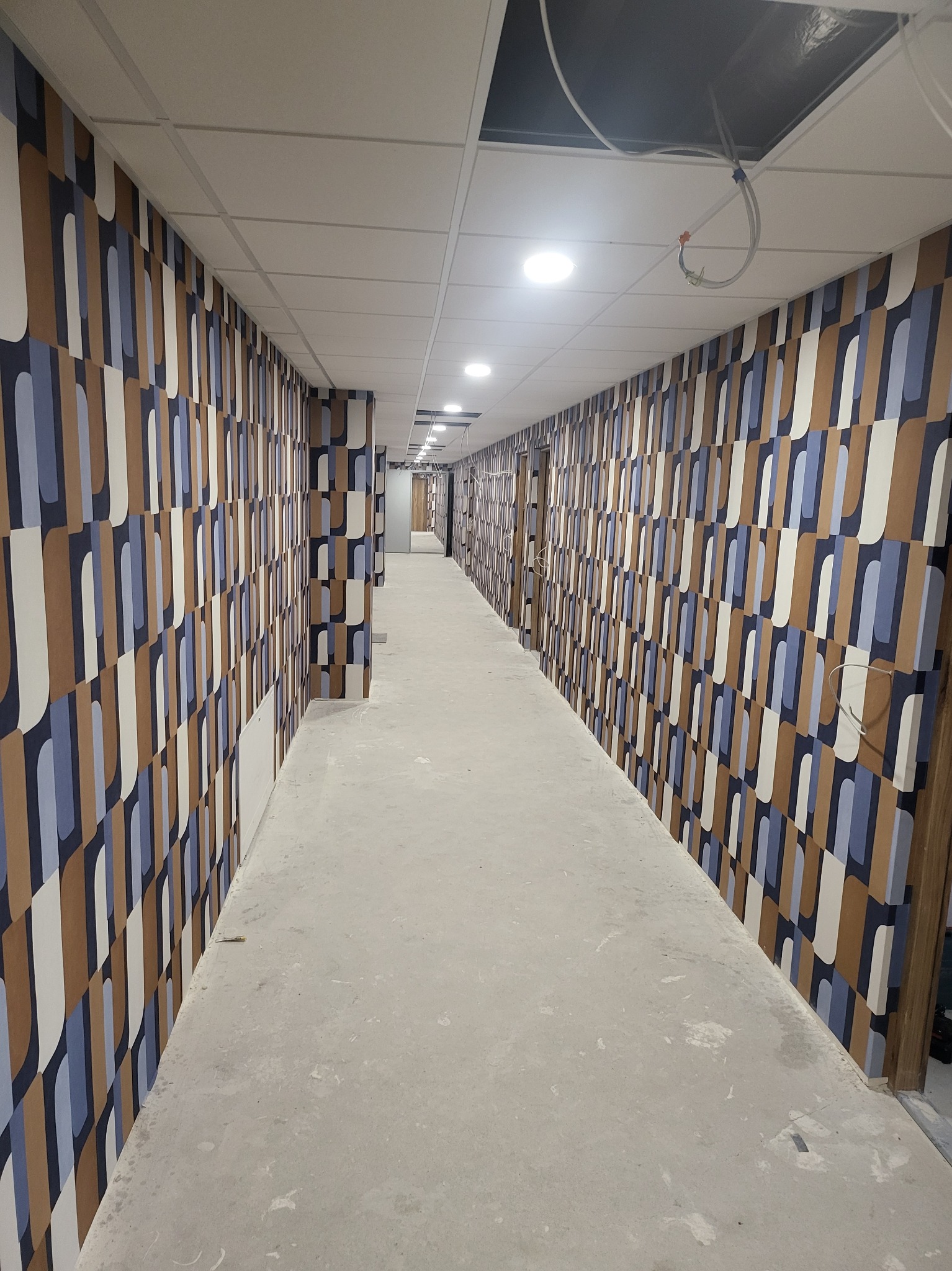 Długi korytarz wykończony tapetą w geometryczne wzory w odcieniach brązu, bieli i błękitu, widok z perspektywy posadzki w trakcie prac wykończeniowych, z widocznymi śladami po budowie.
