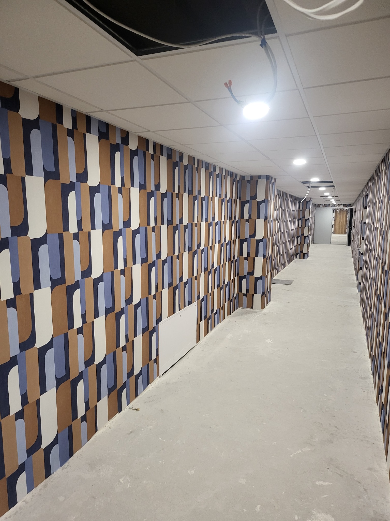 Długi korytarz biurowy z nowo położoną tapetą w geometryczne wzory w odcieniach brązu, błękitu i bieli, sufit podwieszany z widocznymi przewodami. Podłoga surowa, w trakcie wykończenia.