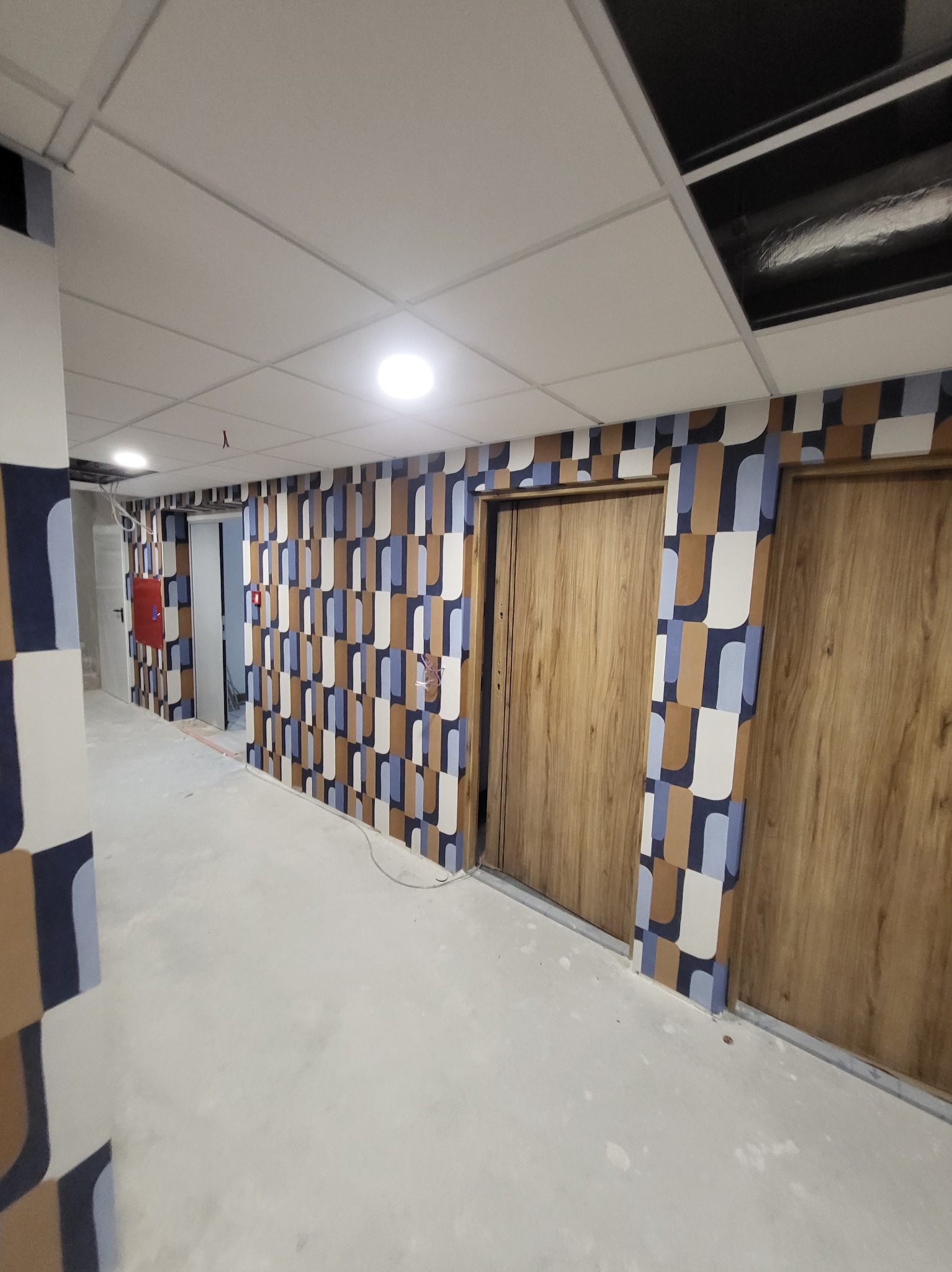 Korytarz biurowy z nową tapetą w geometryczne wzory, widoczne drzwi z drewnianą okleiną i sufit podwieszany.