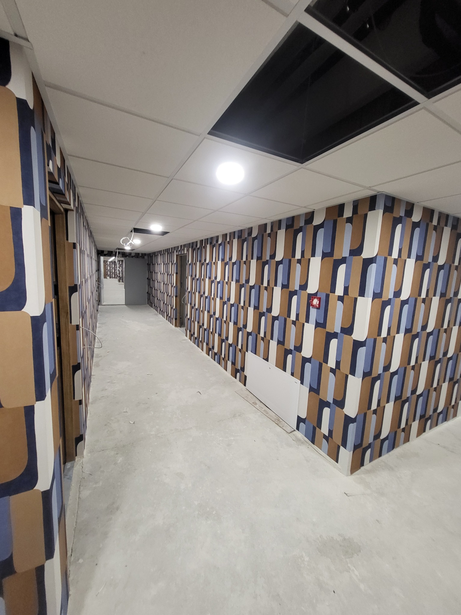 Długi korytarz wykończony tapetą w geometryczne wzory w odcieniach brązu, błękitu i bieli, z widocznymi drzwiami i surowym betonowym podłożem, oświetlony lampami sufitowymi.