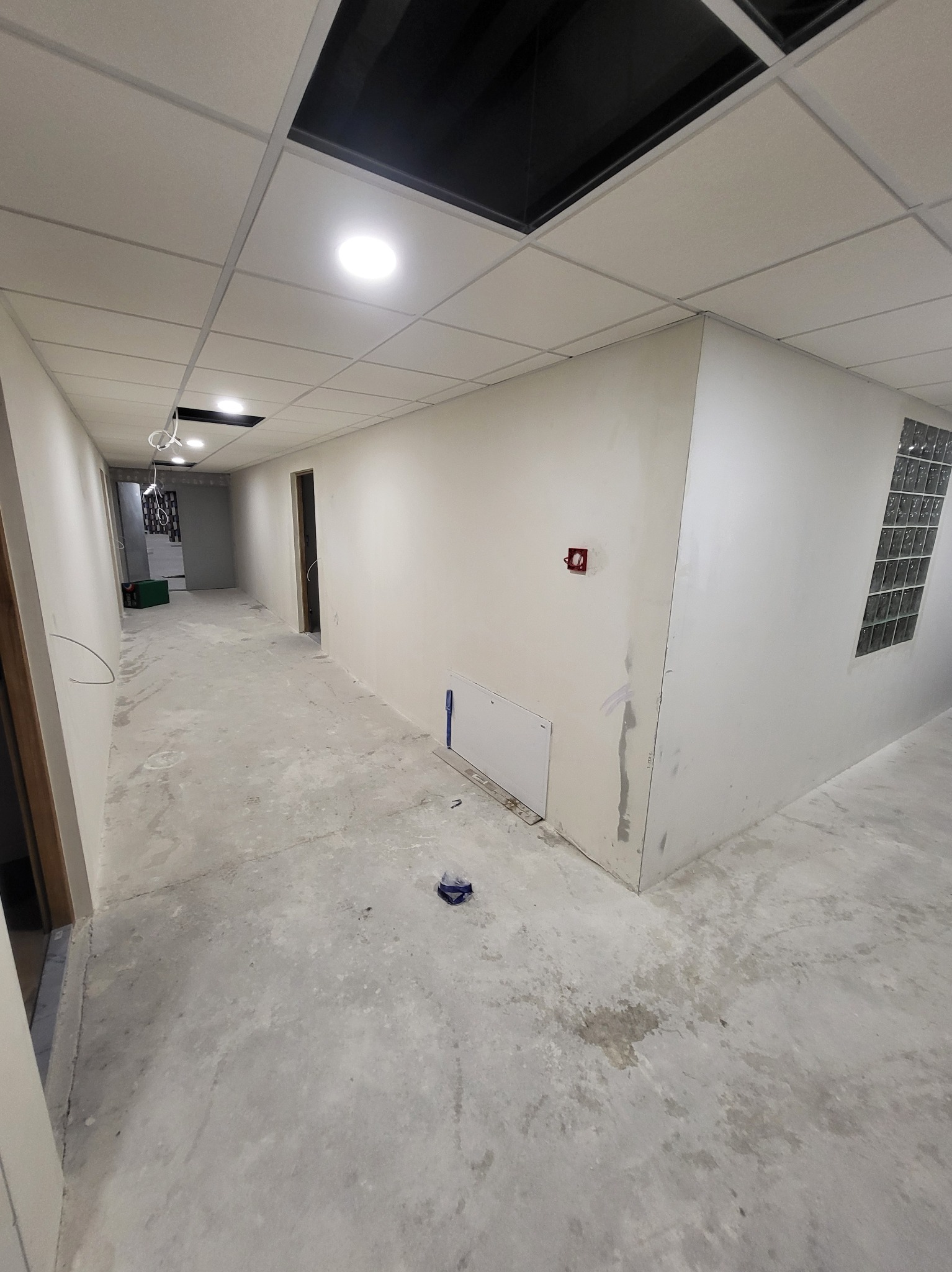 Długi korytarz w trakcie remontu z surowymi ścianami przygotowanymi pod malowanie, widoczny fragment instalacji elektrycznej, sufit podwieszany z otworami, betonowa posadzka z plamami, mała biała...
