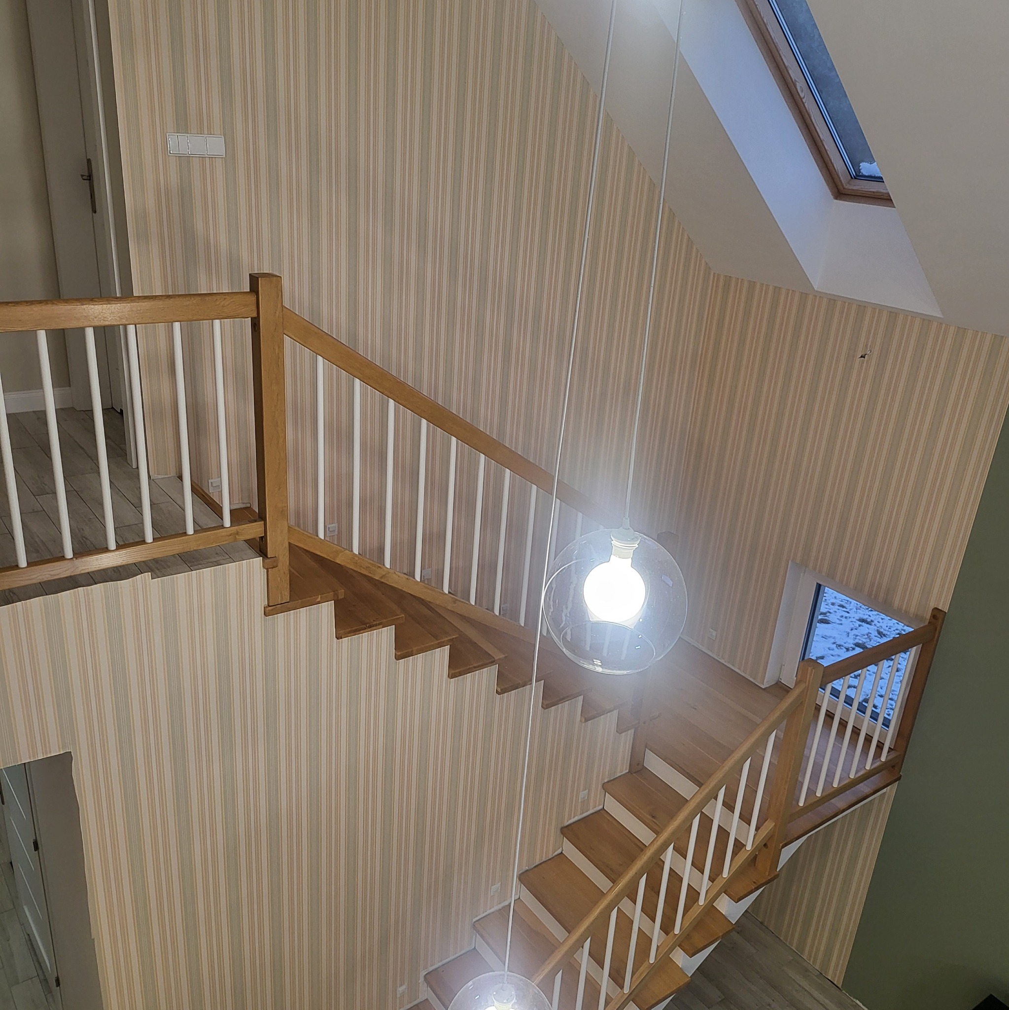 Wnętrze domu po remoncie: schody z drewnianą balustradą i tapetą w pionowe pasy. Widok z góry, lampa wisząca. Okno dachowe i okno na podeście schodowym.