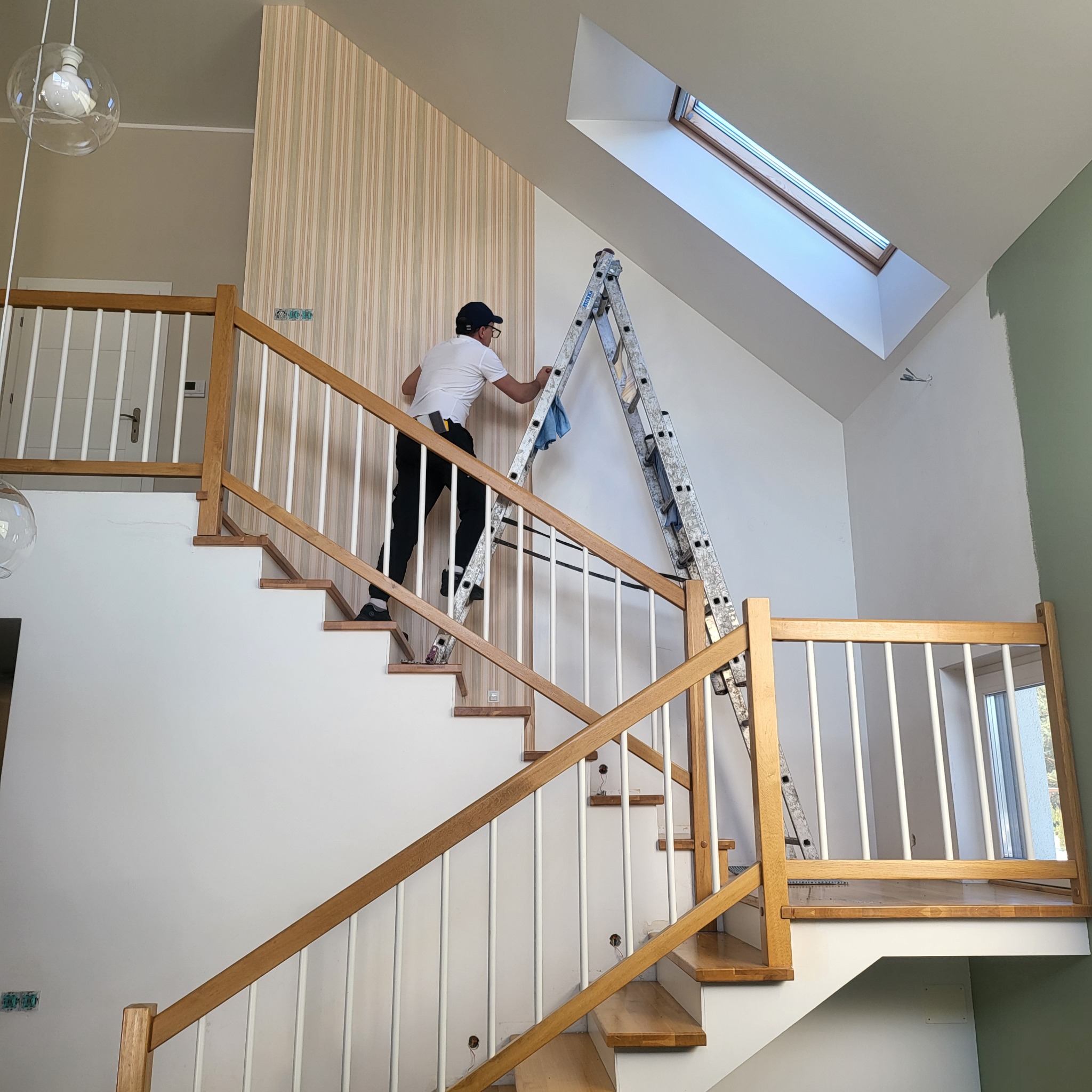 Pracownik na drabinie aplikuje tapetę w paski na wysokiej ścianie klatki schodowej z oknem dachowym. Widoczne drewniane schody z białą balustradą i wisząca lampa.