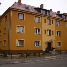 Odnowiona, trzypiętrowa kamienica w ciepłych odcieniach żółci i pomarańczy, z nową elewacją i czerwonym dachem. Widoczne okna z białymi ramami i metalowe rynny.