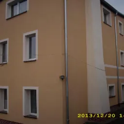 Ocieplona elewacja budynku z oknami w białych ramach i rurą spustową, widok z dołu. Jasna, beżowa kolorystyka. Data wykonania zdjęcia widoczna w prawym dolnym rogu.