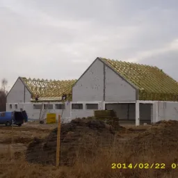 Budowa hali: widok na konstrukcję dachu z drewnianą więźbą, ściany z białego bloczka i pracujących dekarzy. W tle błękitny bus i sterta materiałów budowlanych.