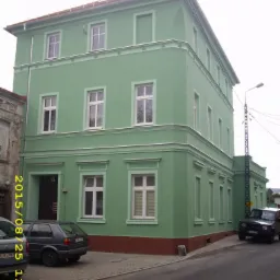 Odnowiona elewacja budynku w Mirsku, pomalowana na pastelowy zielony kolor z białymi obramowaniami okien i ciemnoczerwonym cokołem. Widok na narożnik budynku.