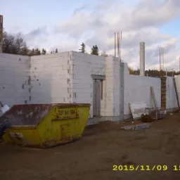 Ściana budynku w trakcie wznoszenia z betonu komórkowego, widoczny kontener na gruz i maszyny budowlane. Częściowe zbrojenie pionowe.