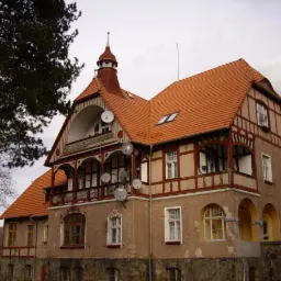 Dwukondygnacyjny budynek z czerwonym dachem i elementami szachulcowymi. Widoczne liczne anteny satelitarne, okna dachowe i kamienna podmurówka.