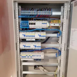 Otwarta szafa rozdzielcza z bezpiecznikami, listwami zaciskowymi i okablowaniem w kolorach niebieskim, ż&oacute;łtym i czarnym, zamontowana na surowej ścianie.