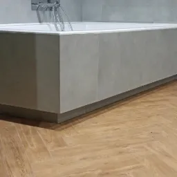 Nowoczesna łazienka z wanną obudowaną szarymi płytkami, chromowaną armaturą i podłogą wyłożoną panelami w jodełkę.