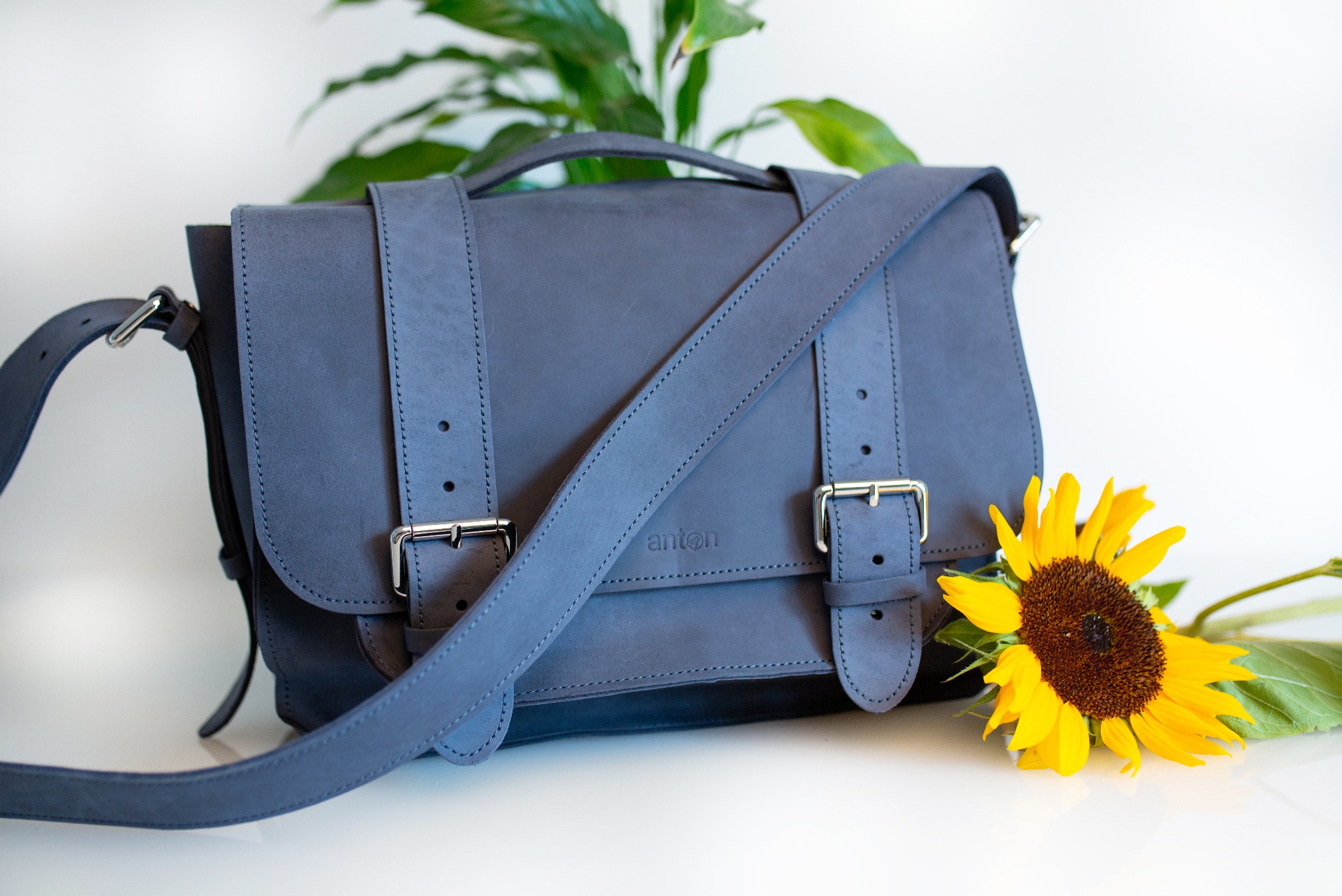 Stylowa listonoszka w odcieniu gołębiej szarości, wykonana ze skóry z subtelnym logo 'anton', leżąca na białym tle obok świeżego słonecznika i zielonej rośliny doniczkowej.