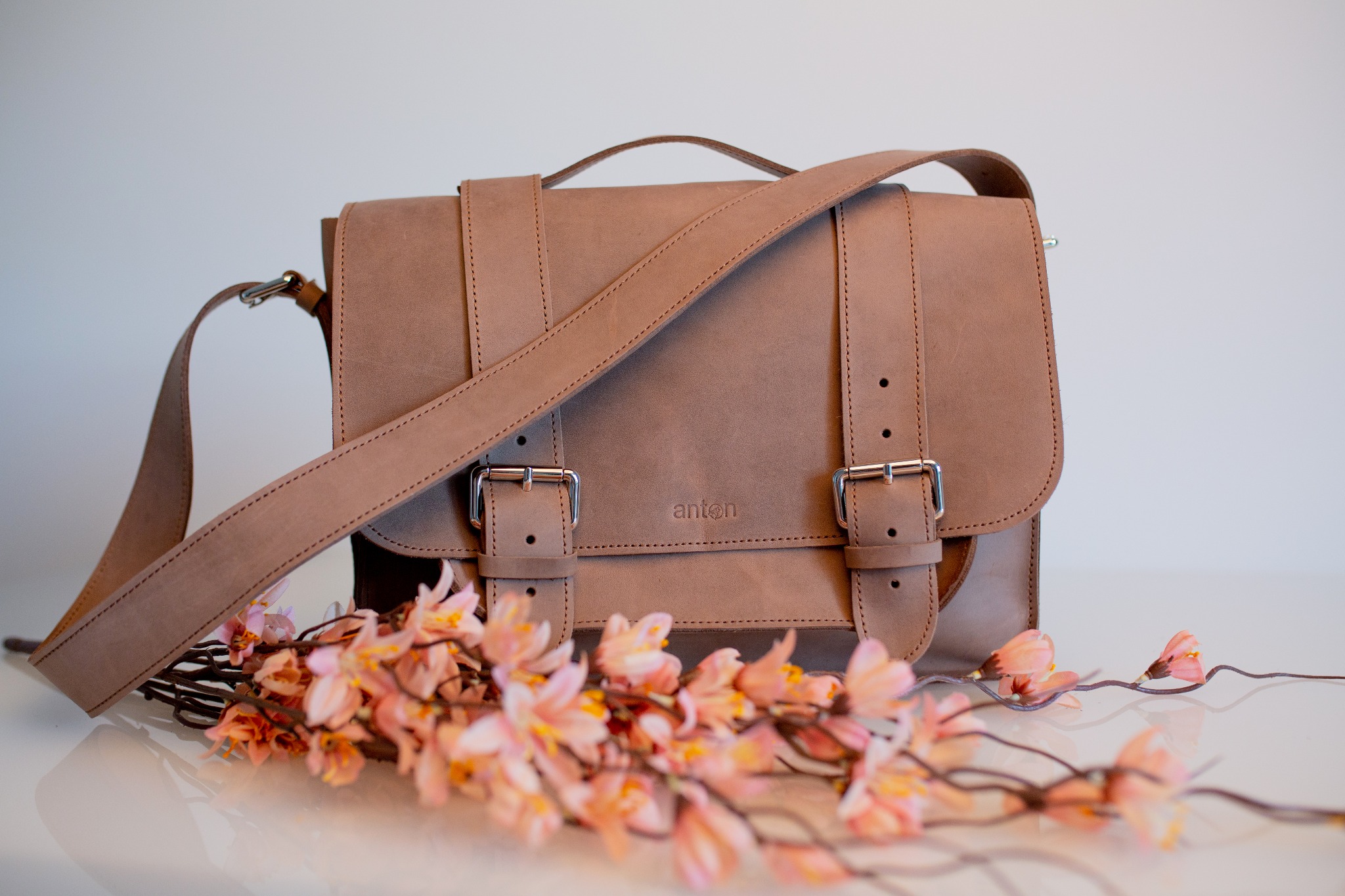 Jasnobrązowa skórzana listonoszka z regulowanym paskiem, zapinana na klamry, z subtelnym logo 'anton' pośrodku, ozdobiona gałązką kwitnących brzoskwiń na błyszczącym blacie.