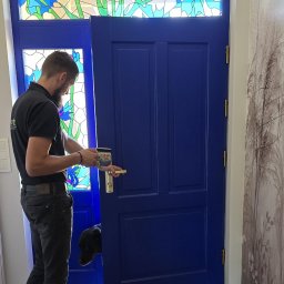 Doorotka - Niebieskie drzwi wejściowe z witrażem kwiatowym nad nimi, otwarte przez mężczyznę trzymającego kubek. Pies zagląda zza drzwi.