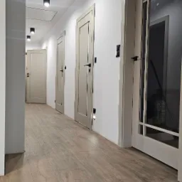 Długi korytarz z drewnianą podłogą, oświetleniem sufitowym i podłogowym. Widoczne drzwi wewnętrzne w odcieniach beżu, z czarnymi klamkami. Jedne z drzwi są szklane.