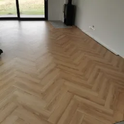 Ułożone panele w jodełkę, jasne drewno, nowoczesny salon z kominkiem i dużym oknem na ogród. Widoczne niedokończone listwy przypodłogowe.