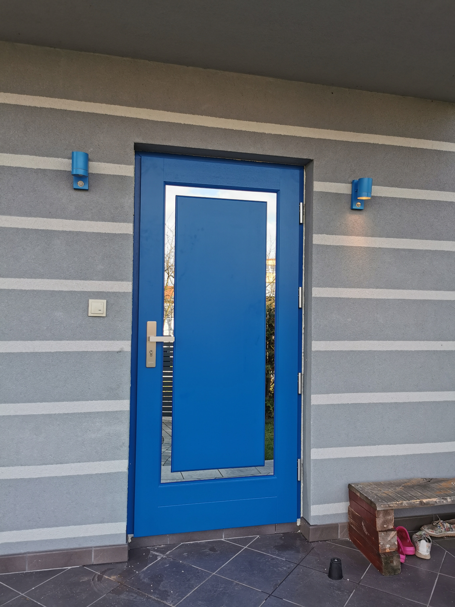 Niebieskie drzwi zewnętrzne z srebrną ramką, zamontowane w szarej ścianie z poziomymi pasami, oświetlone przez dwie niebieskie lampy ścienne.