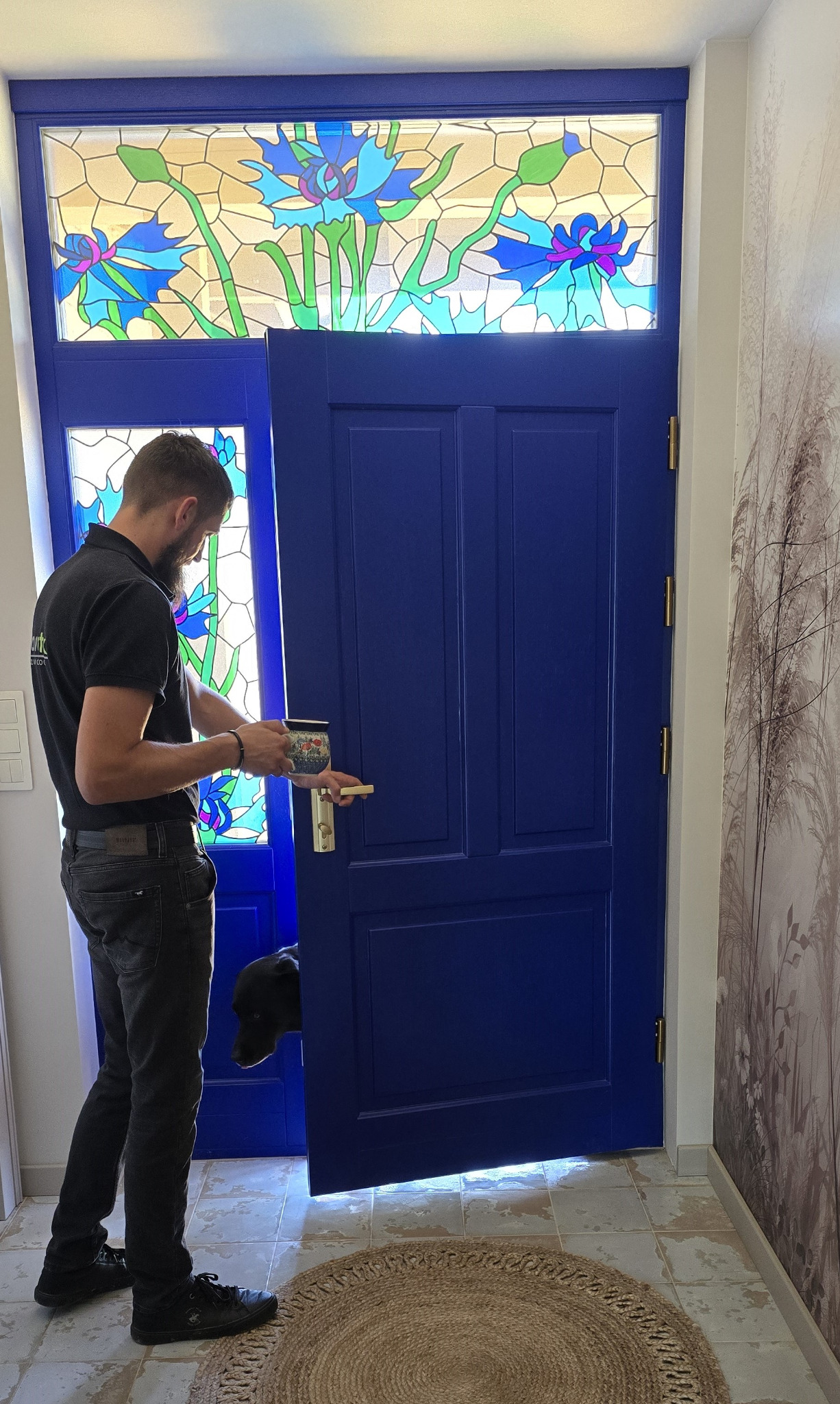 Niebieskie drzwi wejściowe z witrażem kwiatowym nad nimi, otwarte przez mężczyznę trzymającego kubek. Pies zagląda zza drzwi.