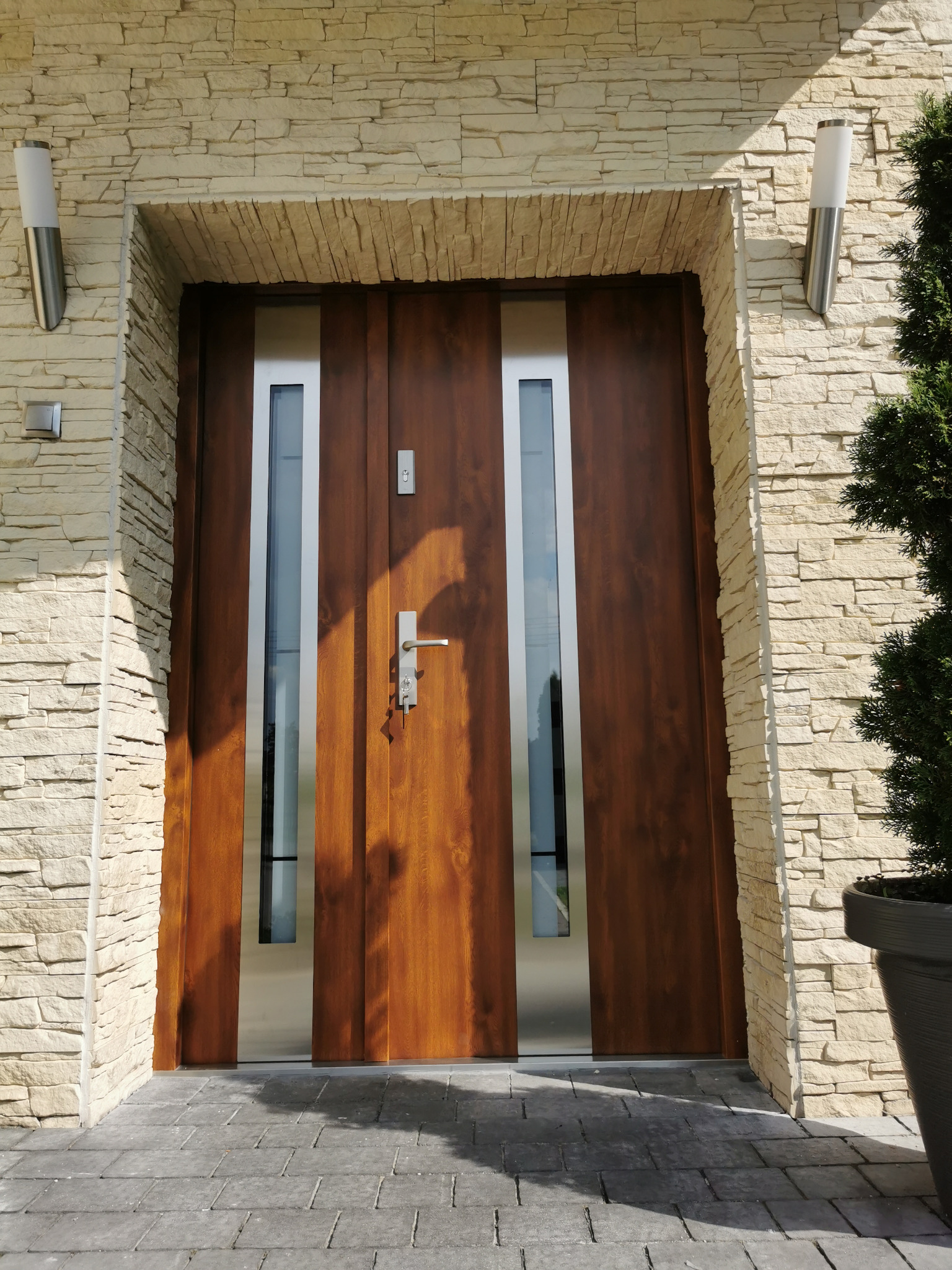 Brązowe drzwi zewnętrzne z szybami w srebrnych ramach, osadzone w elewacji z jasnego kamienia, oświetlone lampami ściennymi i z widoczną rośliną w donicy.