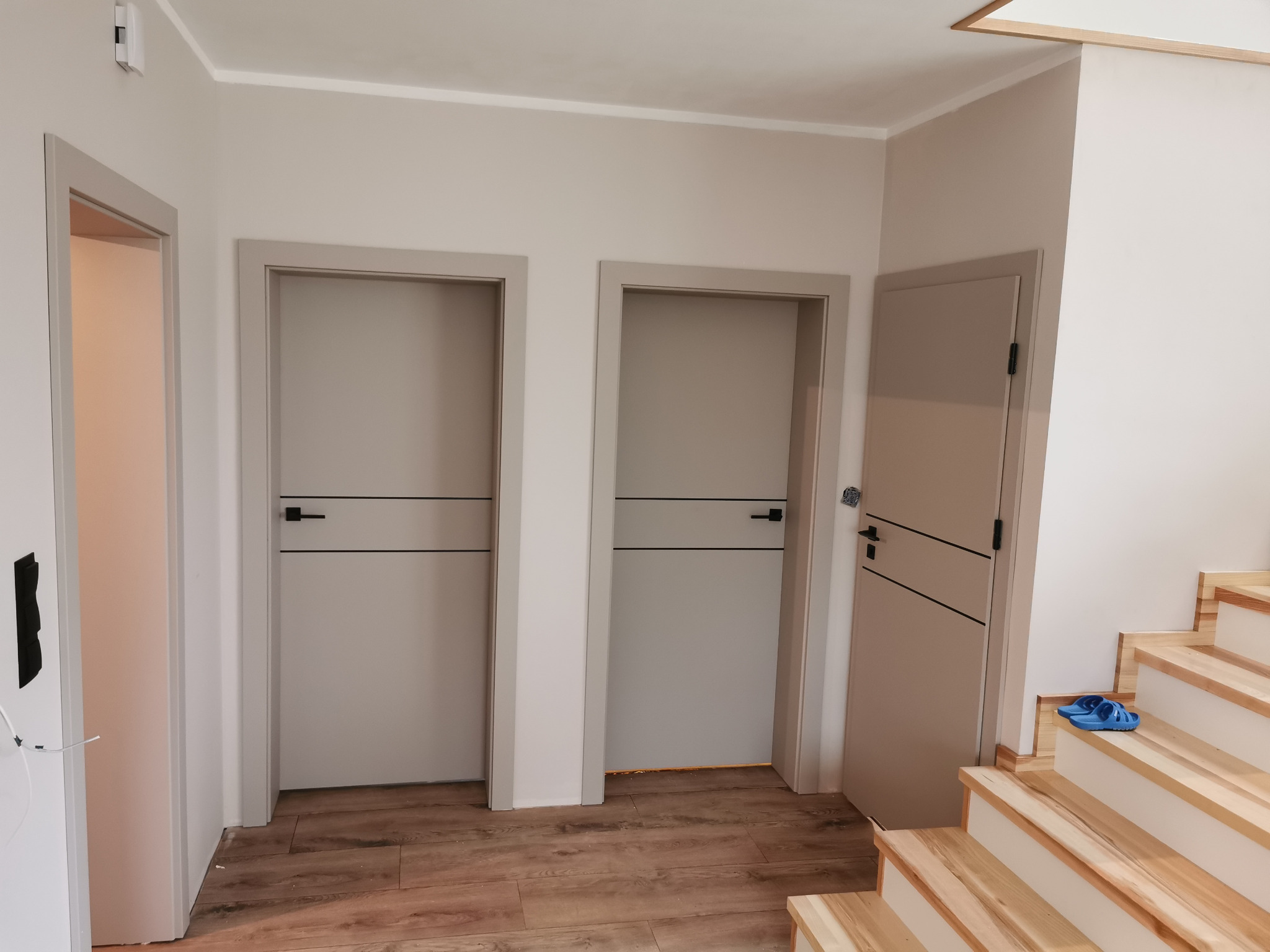 Trzy szare drzwi wewnętrzne z czarnymi poziomymi akcentami w nowym domu. Jedne drzwi otwarte, drewniane schody z boku. Podłoga z paneli.