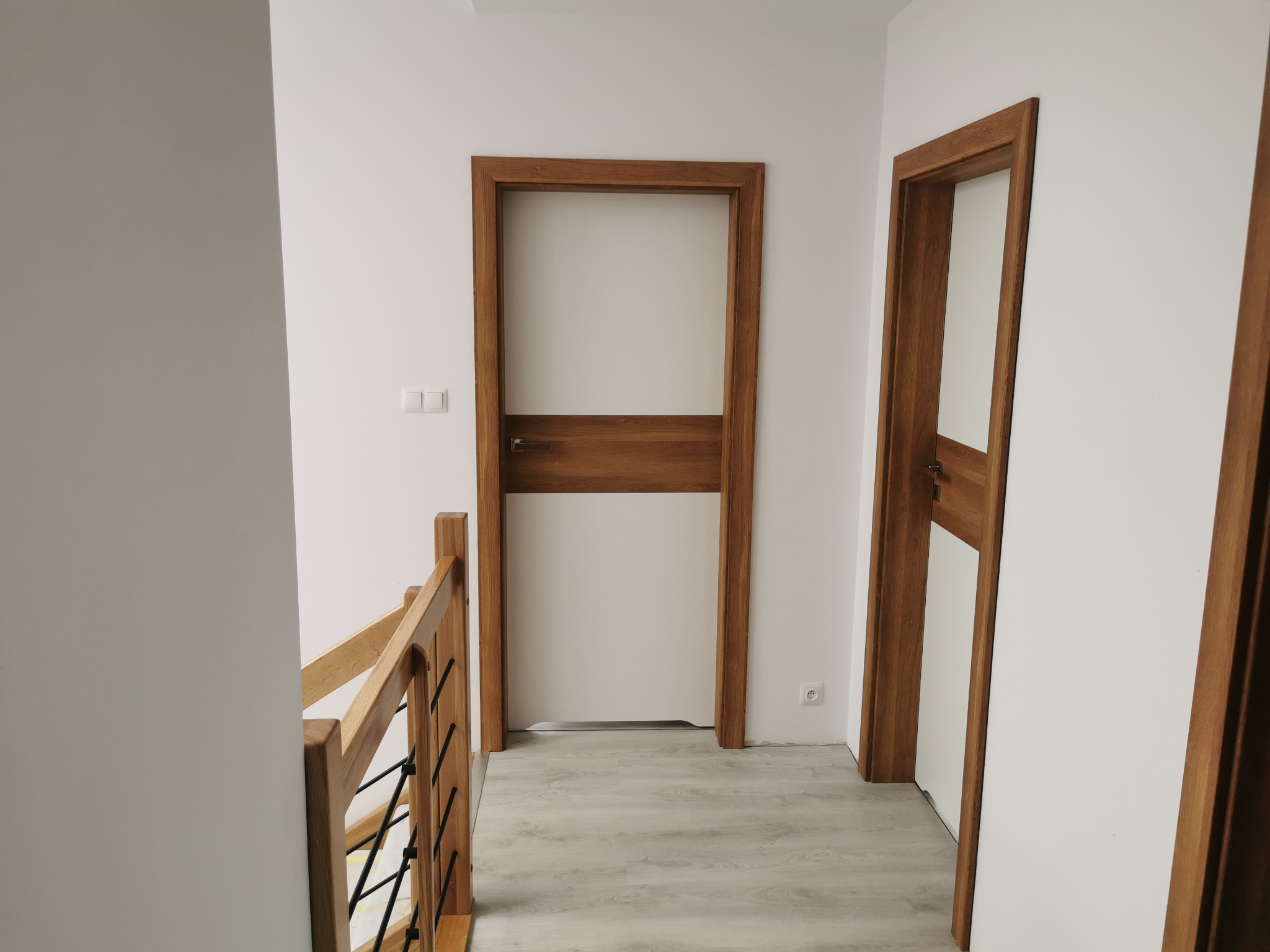 Dwa białe, nowoczesne drzwi wewnętrzne z drewnianą ramą, widoczne z perspektywy klatki schodowej. Jasne wnętrze.