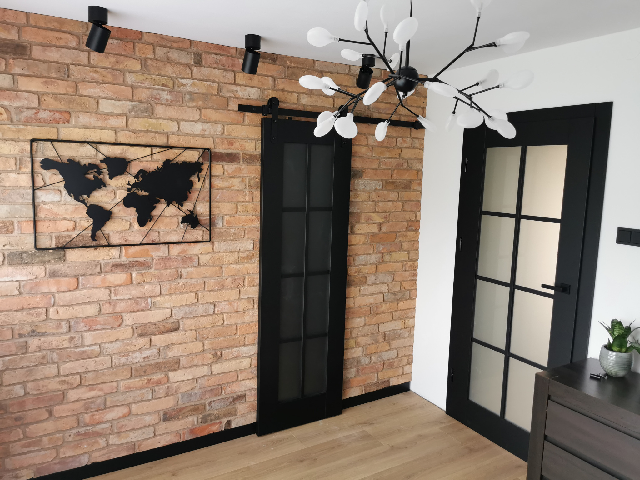 Loftowe wnętrze z ceglaną ścianą, mapą świata i czarnymi drzwiami przesuwnymi z matowym szkłem, oświetlone nowoczesną lampą.