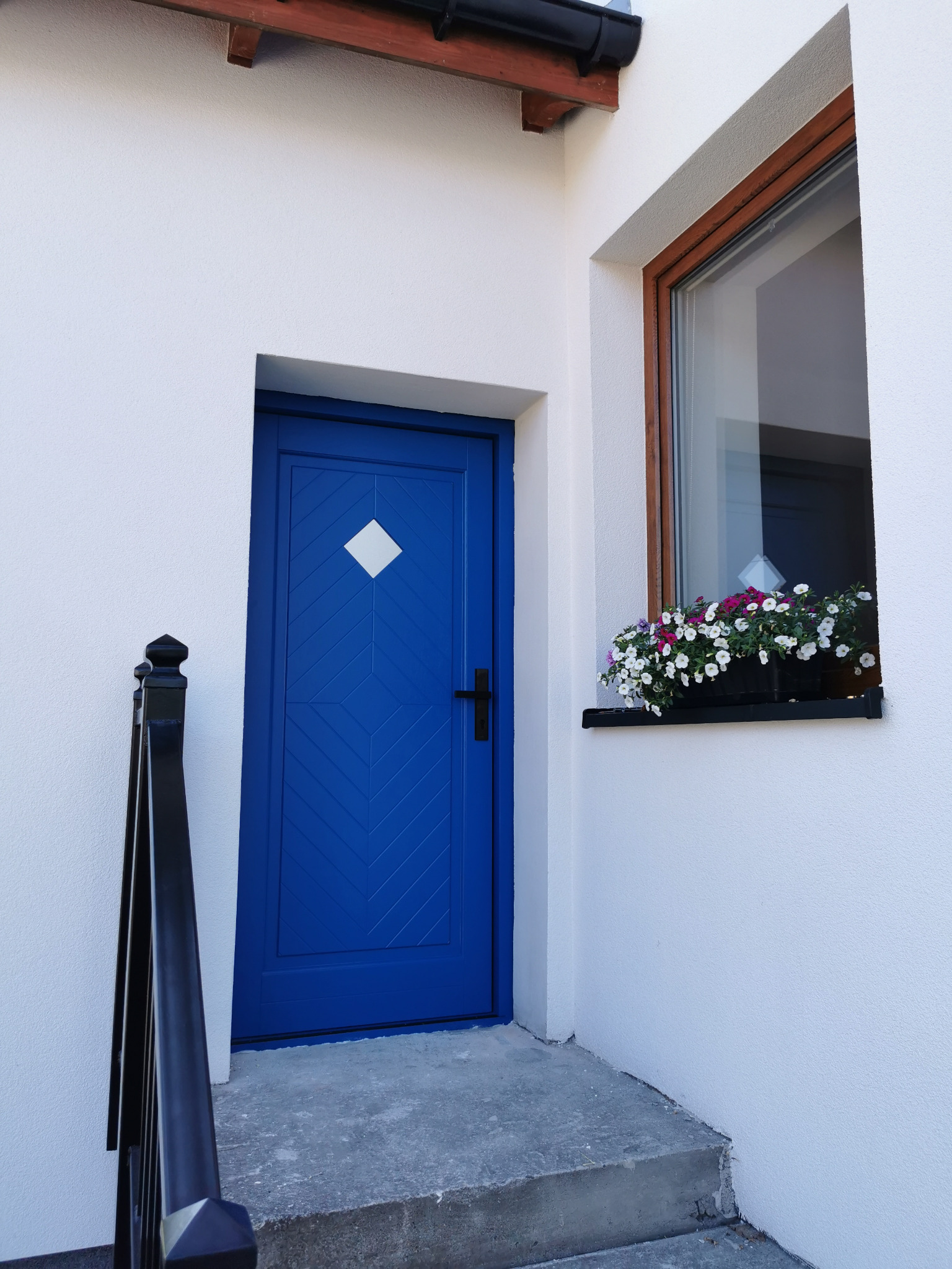 Niebieskie drzwi wejściowe z rombem, okno z kwiatami w doniczce, biała elewacja i schody wejściowe. Ujęcie z dołu.