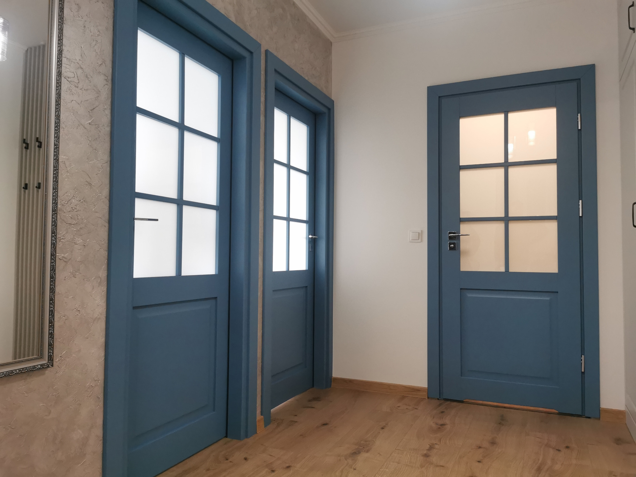 Trzy niebieskie drzwi wewnętrzne z szybami w aranżacji holu. Dwa skrzydła otwarte, trzecie zamknięte. Na ścianie lustro w ozdobnej ramie. Parkiet na podłodze.