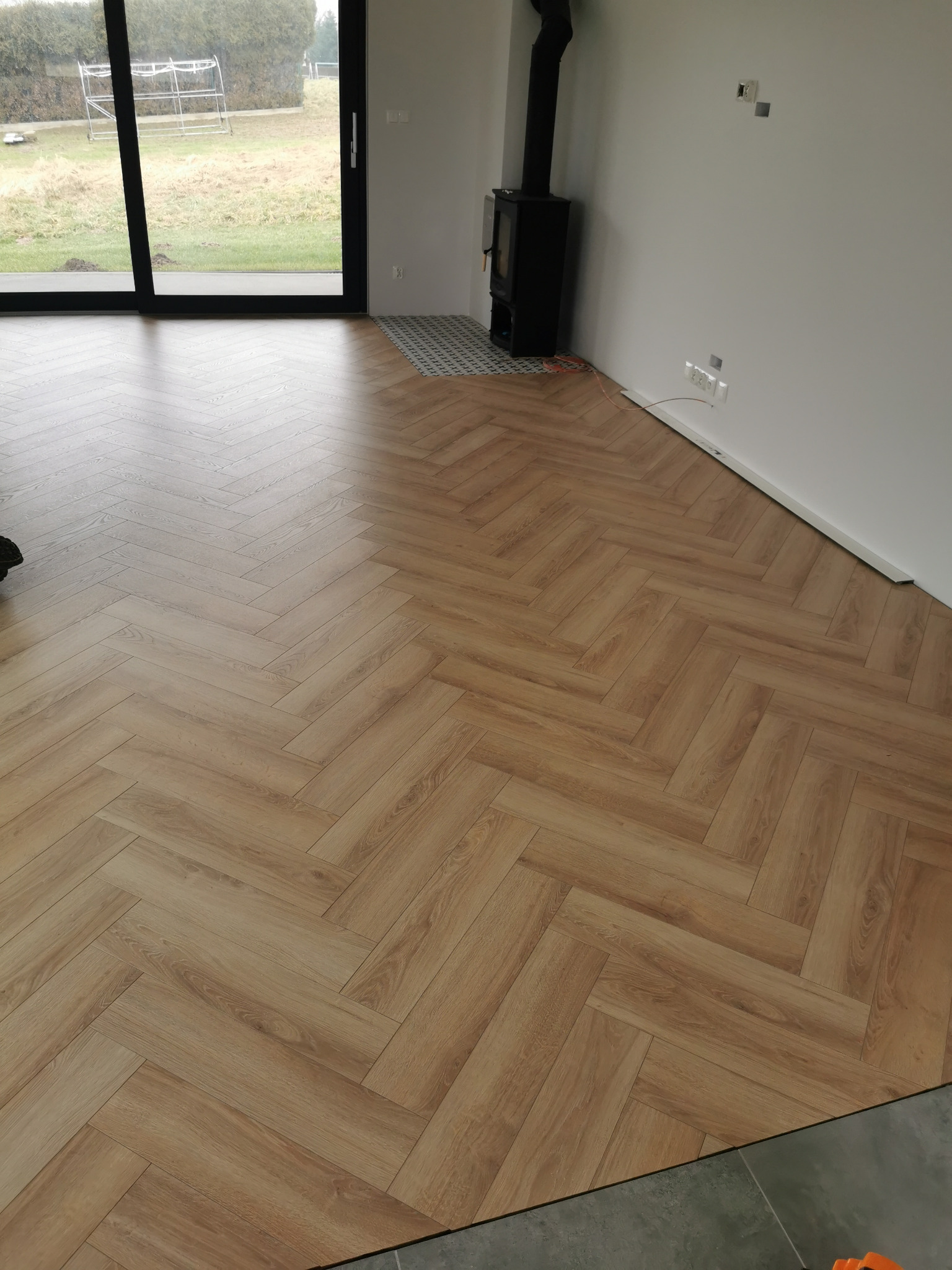 Ułożone panele w jodełkę, jasne drewno, nowoczesny salon z kominkiem i dużym oknem na ogród. Widoczne niedokończone listwy przypodłogowe.