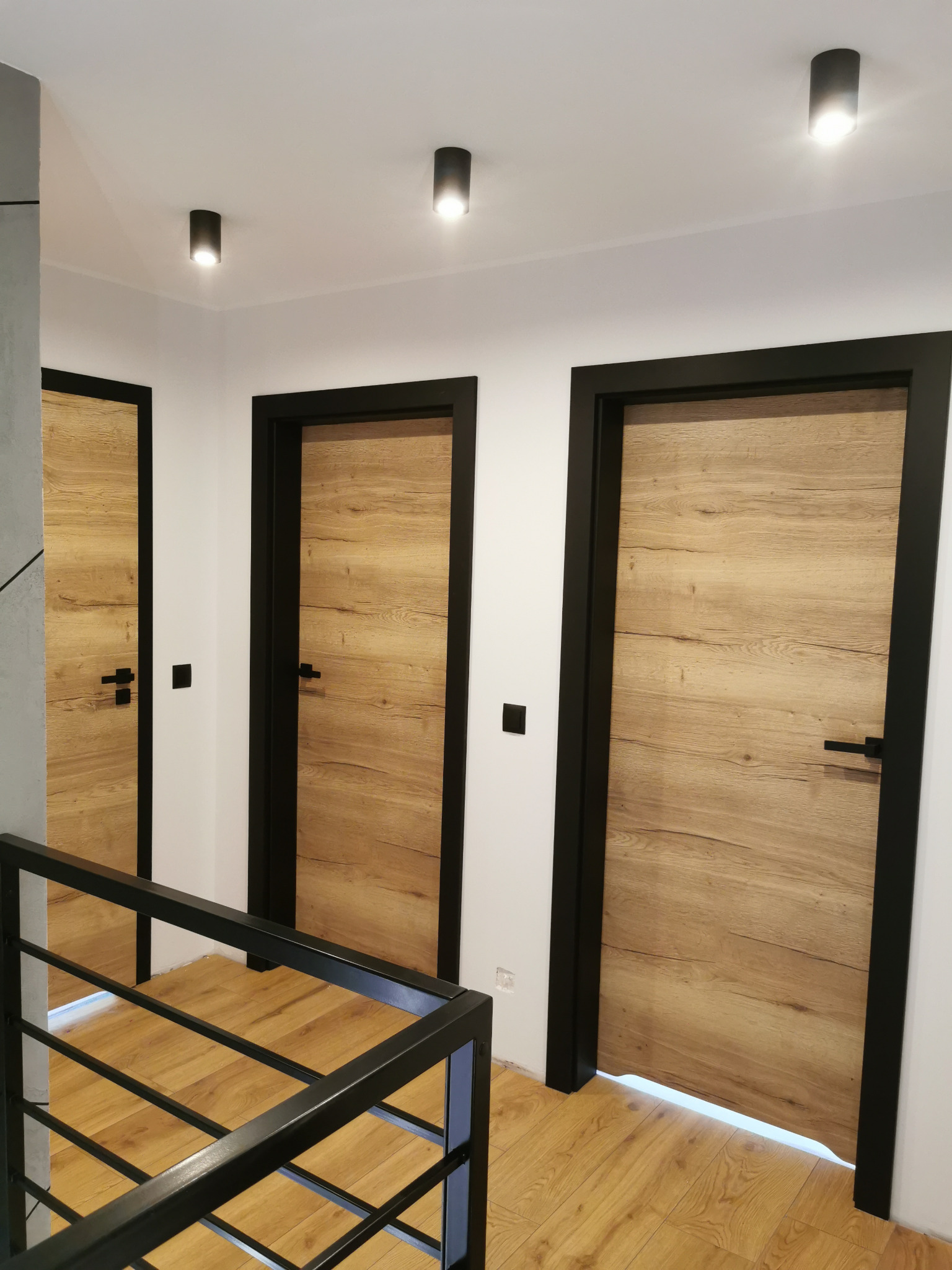 Trzy drewniane drzwi wewnętrzne z czarnymi ościeżnicami i klamkami, widoczne z perspektywy schodów z metalową barierką, oświetlone punktowymi lampami sufitowymi.