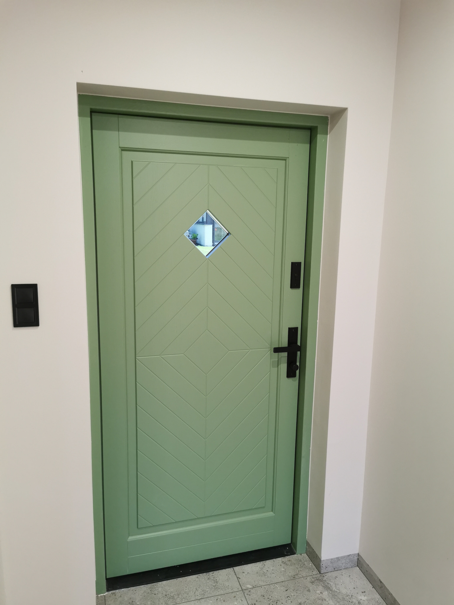 Pistacjowe drzwi z romboidalnym okienkiem i wzorem w jodełkę, czarna klamka, na tle jasnych ścian i szarej podłogi. Widoczny czarny włącznik światła.