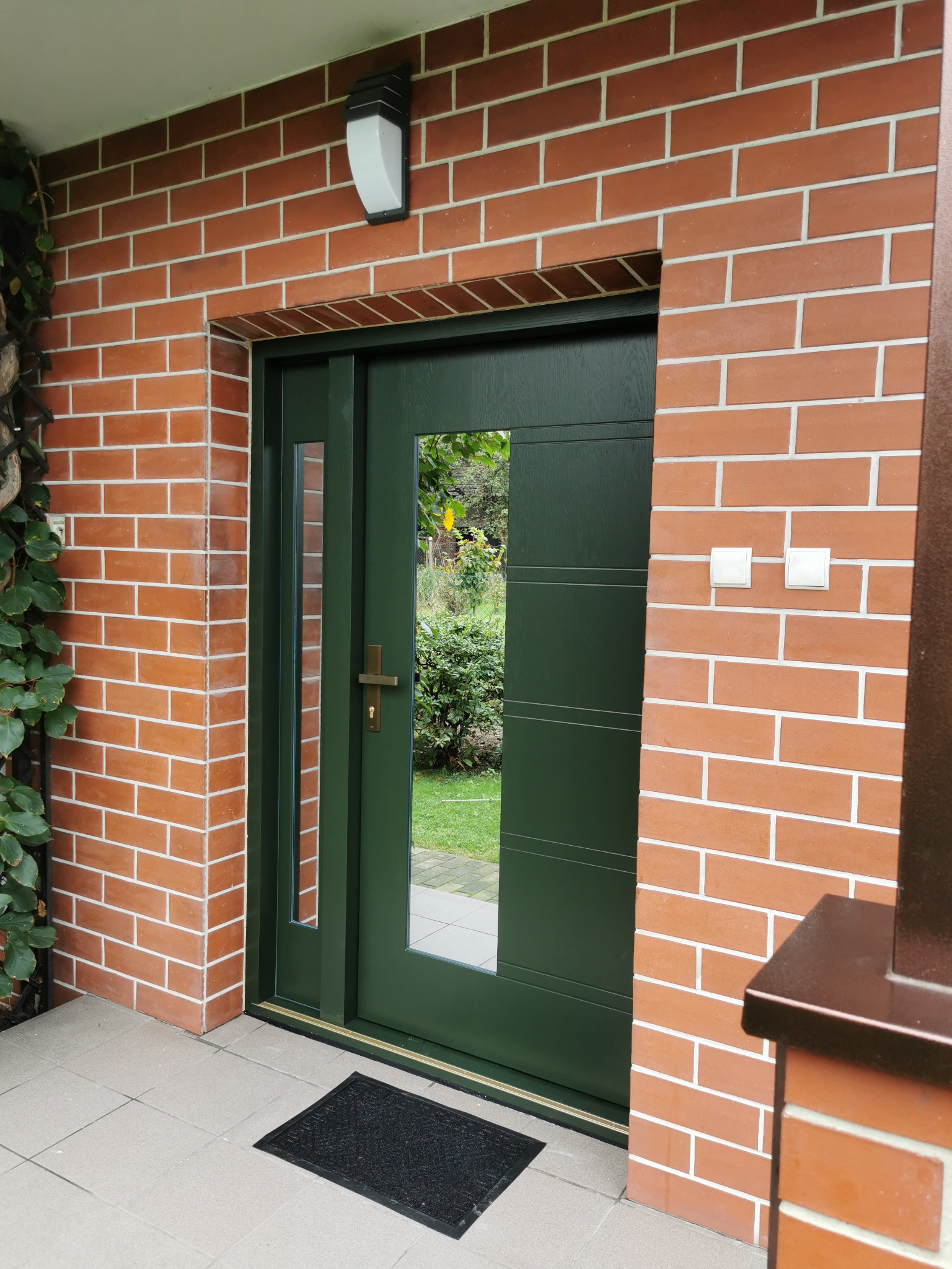 Zielone drzwi wejściowe z szybą w murze z cegły klinkierowej. Widoczne oświetlenie zewnętrzne i wycieraczka. Zewnętrzny widok na ogród przez szybę.