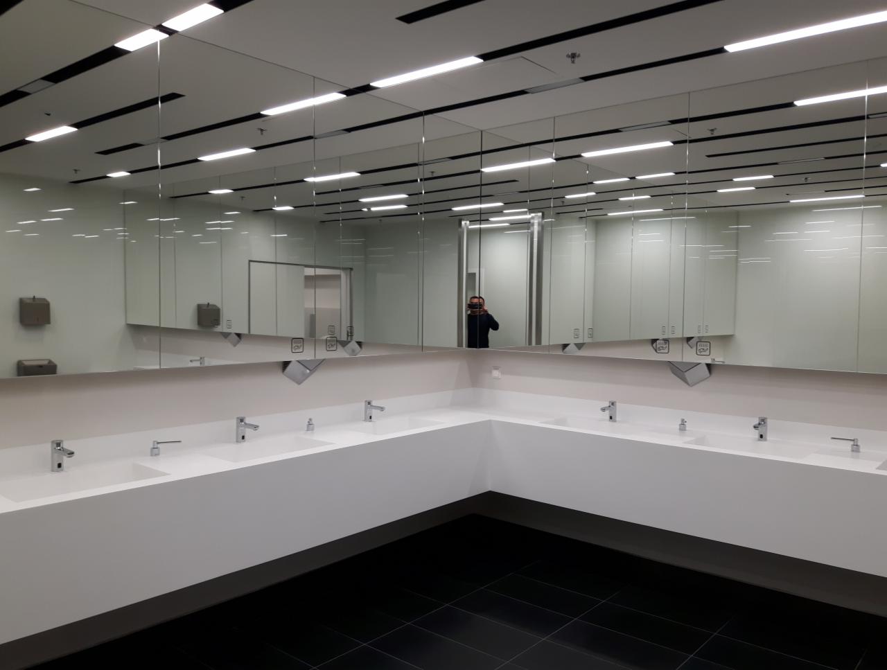Wnętrze czystej, nowoczesnej toalety biurowej z rzędem umywalek w białej zabudowie i dużym lustrem, w którym odbija się osoba robiąca zdjęcie.
