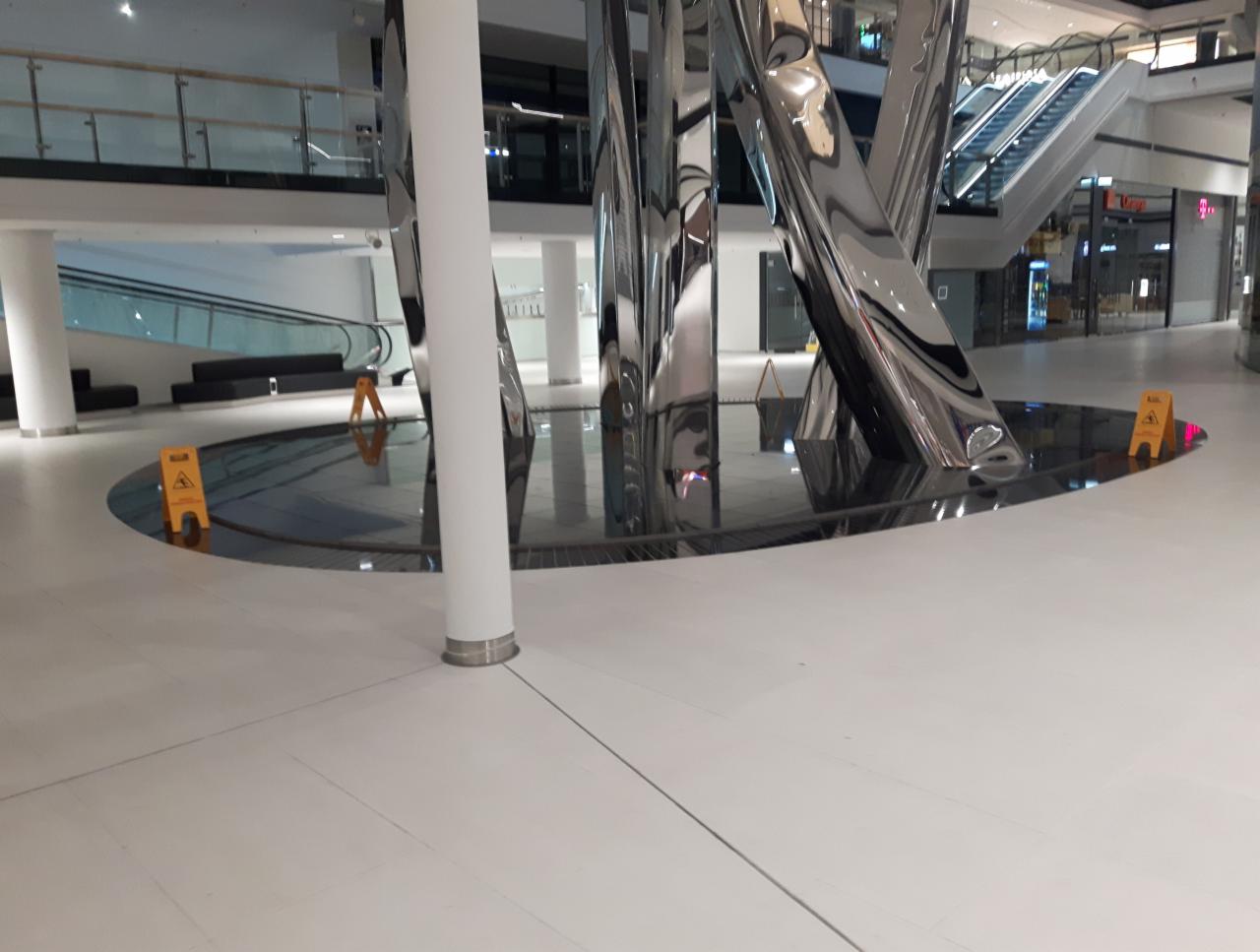 Błyszcząca podłoga w galerii handlowej z nowoczesną fontanną i żółtymi znakami ostrzegawczymi 'Uwaga ślisko'