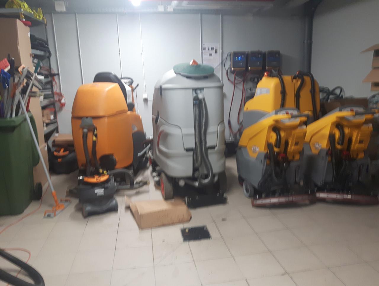 Magazyn z profesjonalnymi maszynami do czyszczenia podłóg: pomarańczowa szorowarka, szara polerka i dwie żółte zamiatarki, widoczne również ładowarki akumulatorów na ścianie.
