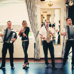 Czwórka muzyków w trakcie występu: dwóch akordeonistów, saksofonista i perkusistka z tamburynem, na tle okna i lustra.