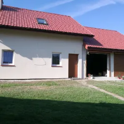 Jasna elewacja domu z czerwoną dachówką, widoczne dwa okna, brązowe drzwi wejściowe oraz brama garażowa z częściowo opuszczoną roletą.