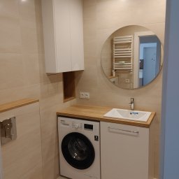 ROBERT DAWID - Nowoczesna łazienka z pralką pod drewnianym blatem, okrągłe lustro odbijające grzejnik drabinkowy, beżowe płytki na ścianach.