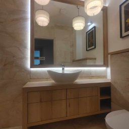 ROBERT DAWID - Jasna łazienka z drewnianą szafką pod umywalką, owalną białą umywalką nablatową, podświetlanym lustrem w drewnianej ramie i trzema wiszącymi lampami z frędzlami.