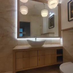 Jasna łazienka z drewnianą szafką pod umywalką, owalną białą umywalką nablatową, podświetlanym lustrem w drewnianej ramie i trzema wiszącymi lampami z frędzlami.