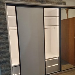 ROBERT DAWID - Biała szafa przesuwna z oświetleniem LED, wbudowana w ścianę z dekoracyjnym kamieniem, widoczna półka z drążkiem i dwie szuflady.